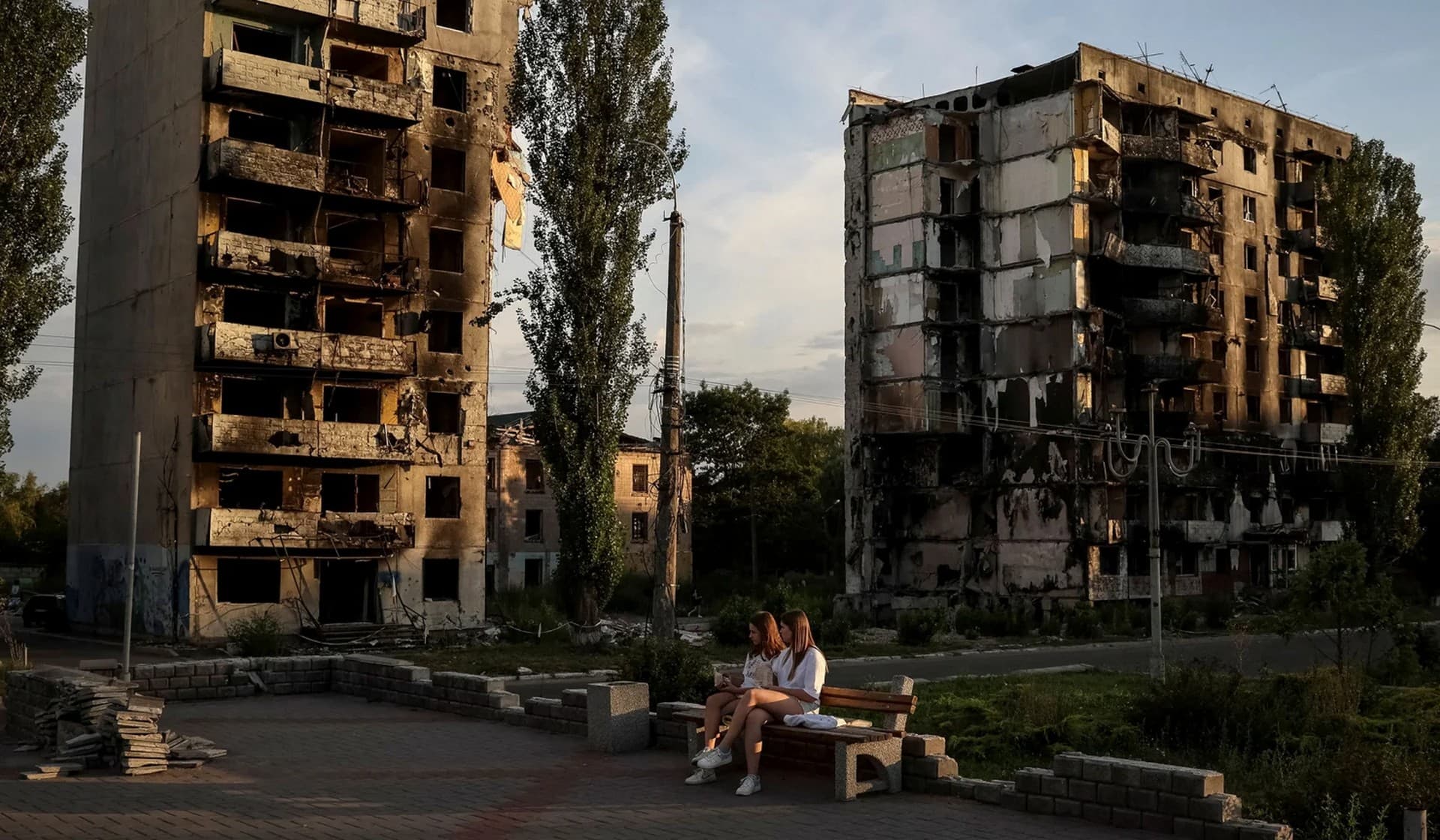 Two girls sit in a public square in front of destroyed buildings in Borodianka
