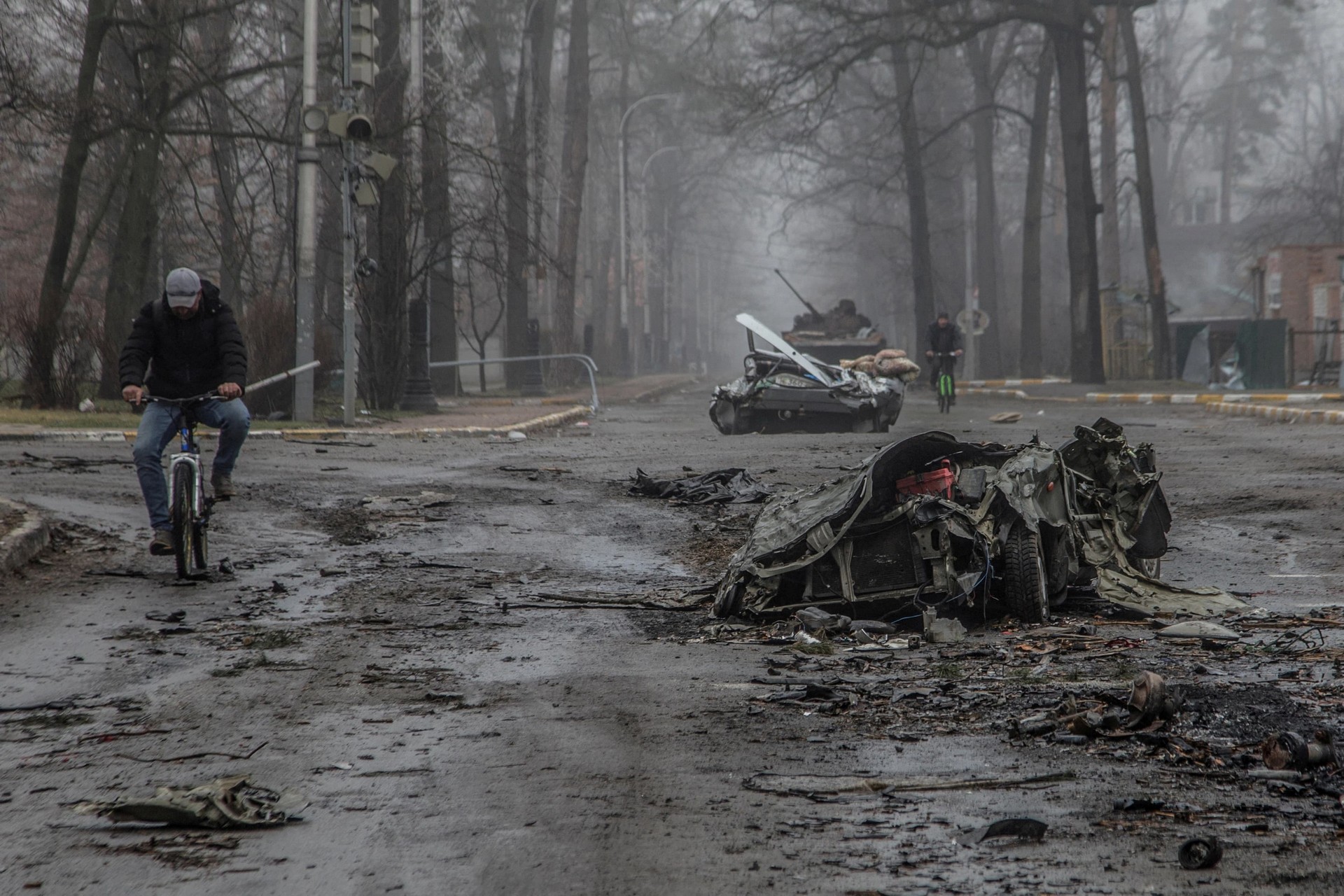 Local residents ride bicycles past flattened civilian cars in the town of Bucha
