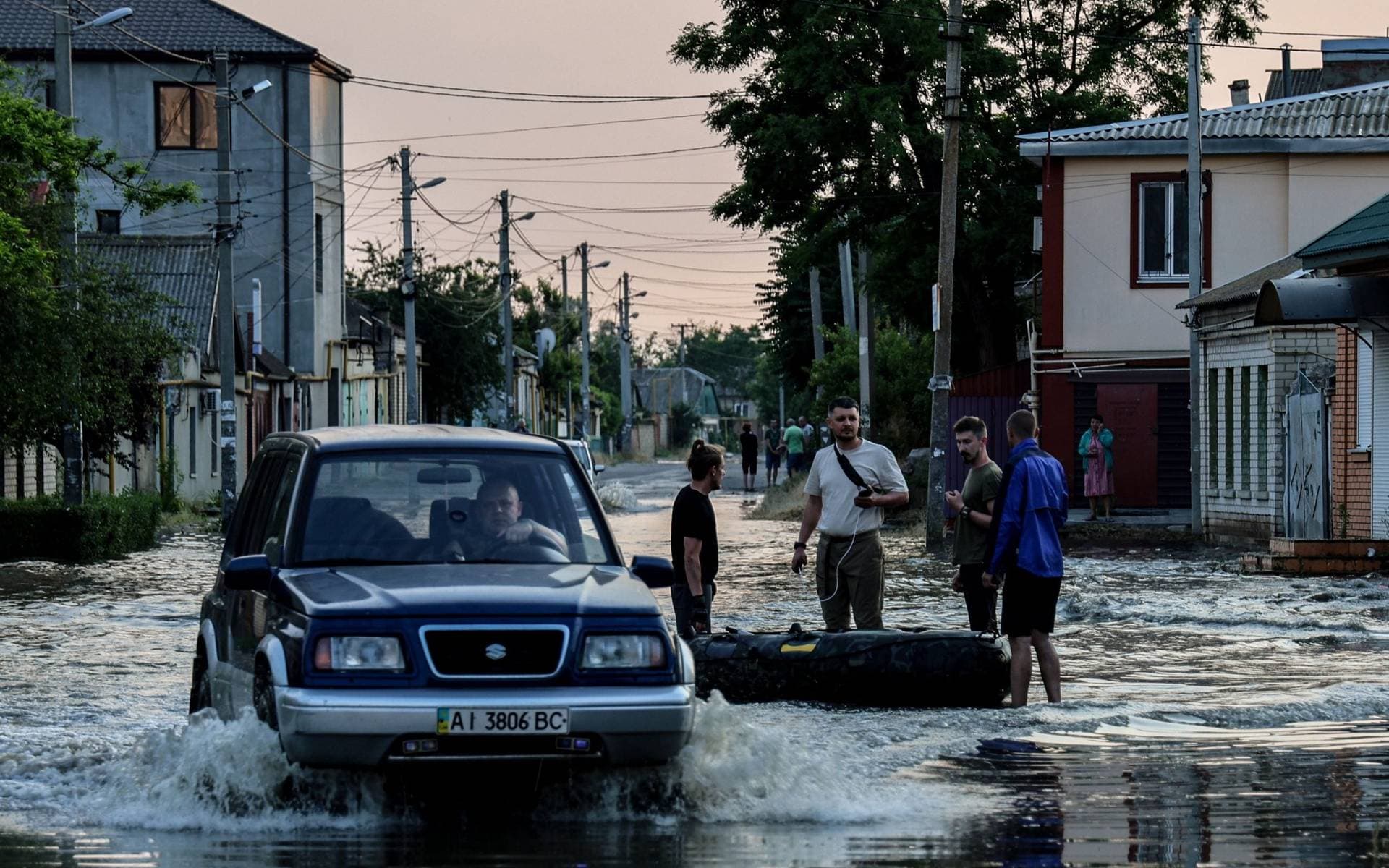 A car makes its way past people standing next to an inflatable boat, in a flooded street of Kherson