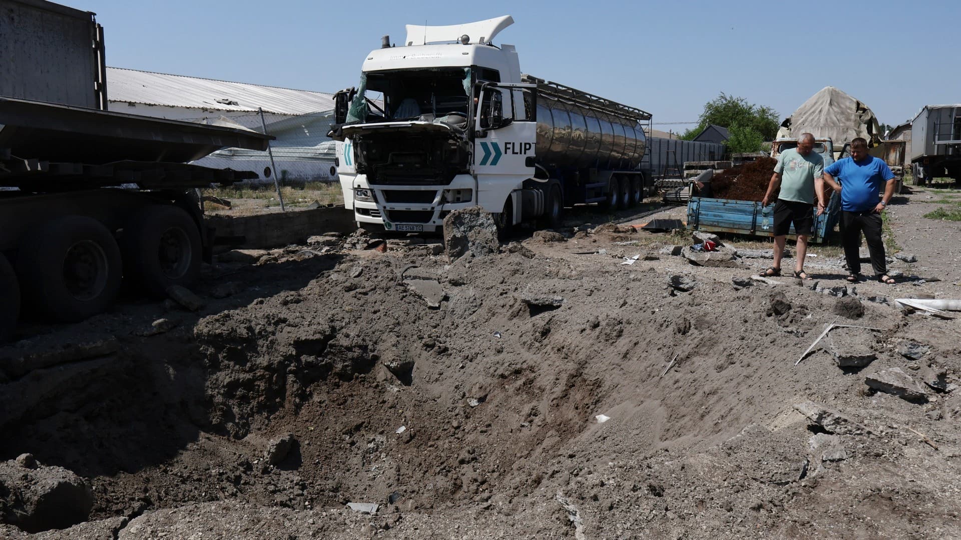 Men look at a crater next to destroyed trucks after Russian shelling in Rozumivka